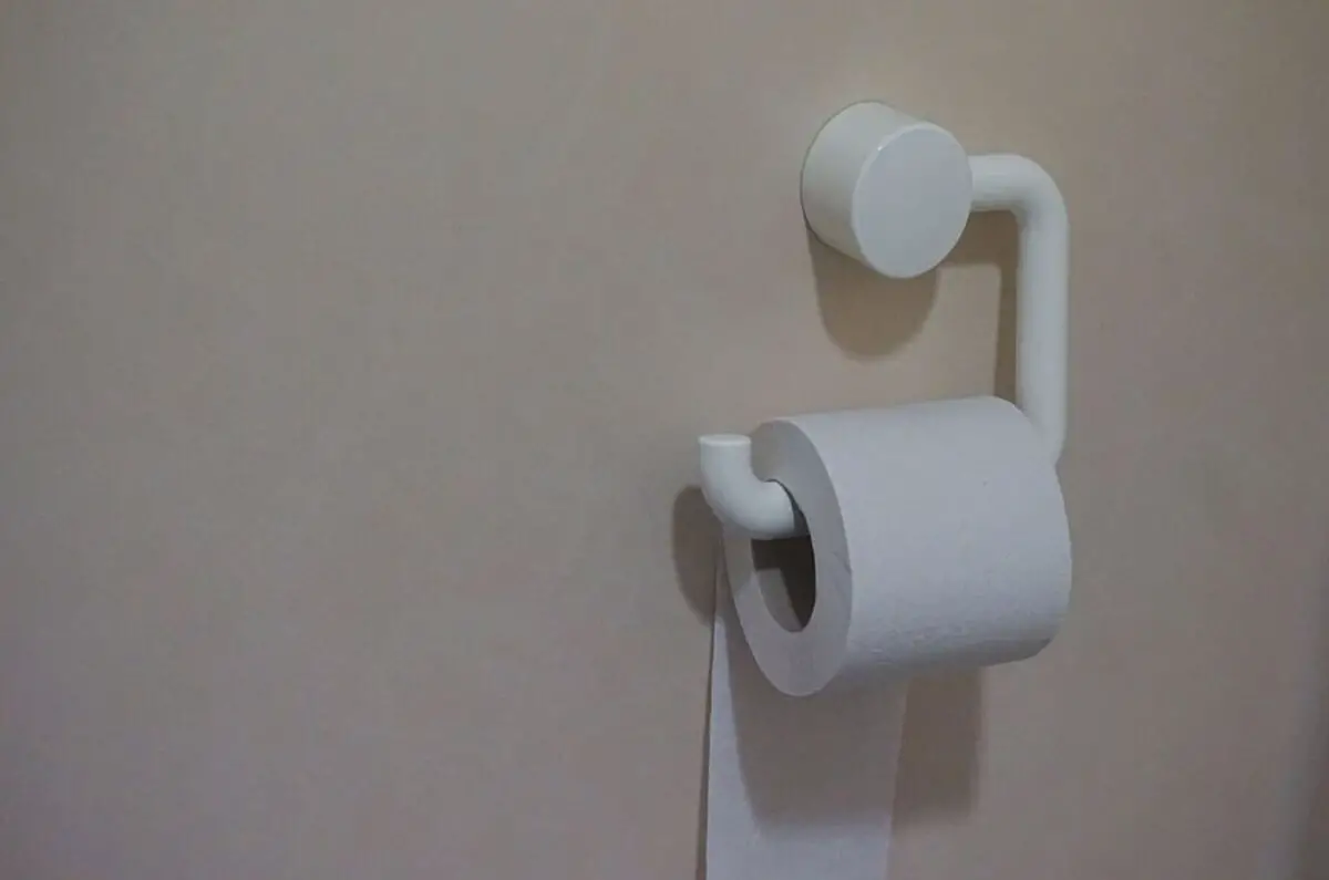 White roll of toilet paper attached on a white toilet paper holder that is installed on a nude colored bathroom wall